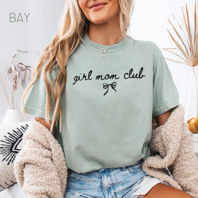 Peut inclure: Un t-shirt vert clair avec le texte "girl mom club" en &eacute;criture noire avec un n&oelig;ud noir en dessous.