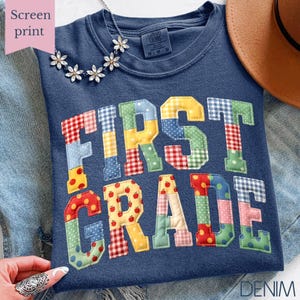 May include: A navy blue t-shirt with the words "FIRST GRADE" in colorful patchwork letters. The shirt has a crew neck and short sleeves. A silver necklace with flower-shaped pendants is draped on the shirt.