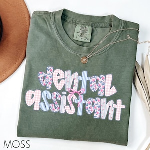 Pode incluir: Camiseta verde musgo com as palavras "dental assistant" em um design floral e xadrez. Um colar de ouro com um pingente est&aacute; sobre a camisa. A camisa tem uma etiqueta Comfort Colors.