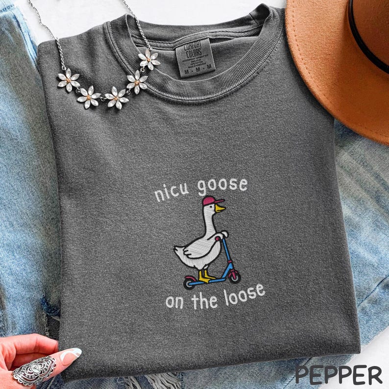 Peut inclure: T-shirt gris fonc&eacute; avec la phrase brod&eacute;e "nicu goose on the loose" et un dessin d'oie sur un scooter. L'oie porte une casquette rose. Un collier de fleurs argent&eacute; et un chapeau marron sont &eacute;galement visibles.
