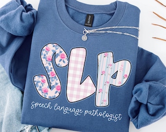 May include: Indigo blue sweatshirt featuring the letters "SLP" in floral, gingham, and striped patterns. The words "speech language pathologist" are written below. A silver necklace with a small pendant is draped across the sweatshirt.
