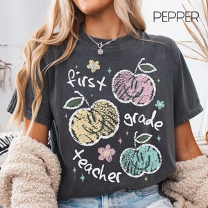 May include: A charcoal gray t-shirt featuring a graphic print with the text "first grade teacher" and "PEPPER". The design includes three textured, colorful apples with smiley faces in pink, yellow, and teal, accented by small flowers and stars.