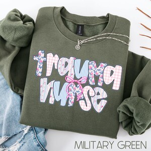 Puede incluir: Sudadera verde militar con la frase "Trauma Nurse" en un dise&ntilde;o floral y de cuadros. Un collar plateado con un colgante de sol est&aacute; sobre la sudadera. La sudadera est&aacute; doblada sobre unos vaqueros azules.