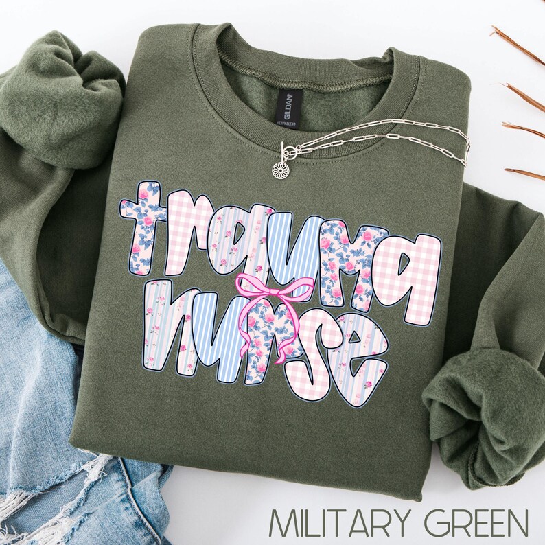 Puede incluir: Sudadera verde militar con la frase "Trauma Nurse" en un dise&ntilde;o floral y de cuadros. Un collar plateado con un colgante de sol est&aacute; sobre la sudadera. La sudadera est&aacute; doblada sobre unos vaqueros azules.