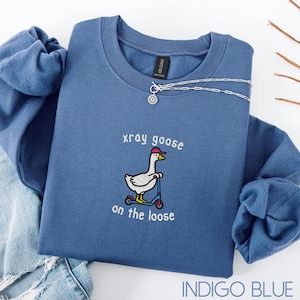 K&ouml;nnte beinhalten: Indigoblaues Sweatshirt mit dem gestickten Schriftzug "xray goose on the loose" und einer Cartoon-Gans auf einem Roller, die eine rote Kappe tr&auml;gt. Eine silberne Halskette mit Sonnenanh&auml;nger ist auf dem Sweatshirt.