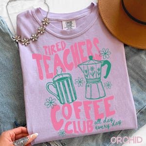 May include: A light purple Comfort Colors t-shirt with a pink and green graphic. The graphic says "Tired Teachers Coffee Club" with images of a coffee mug and a coffee pot. The words "all day every day" are also included.
