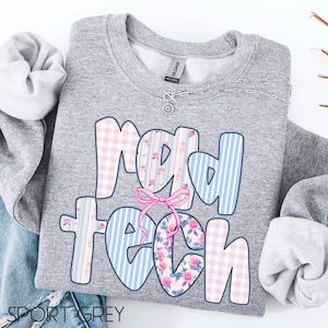 May include: A heather gray sweatshirt with the words "rad tech" in a colorful design. The letters are decorated with pink gingham, blue stripes, and floral patterns. A pink bow is centered between the words. A silver necklace is visible.