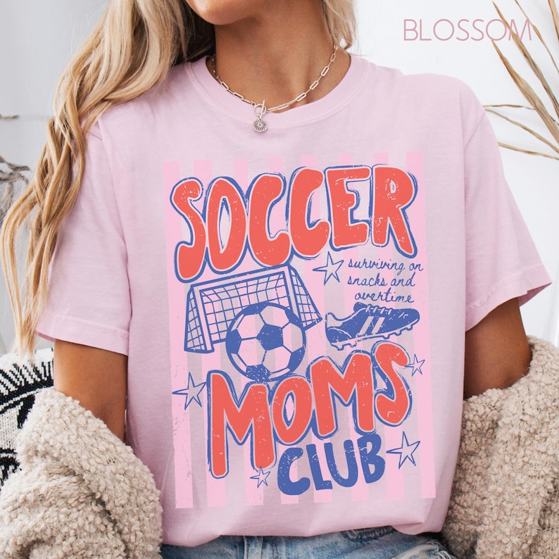 Puede incluir: Camiseta rosa claro con un dise&ntilde;o gr&aacute;fico desgastado. El dise&ntilde;o presenta las palabras "SOCCER MOMS CLUB" en rojo y azul, junto con im&aacute;genes relacionadas con el f&uacute;tbol, como un bal&oacute;n, una porter&iacute;a y botas. Tambi&eacute;n incluye el texto "surviving on snacks and overtime".