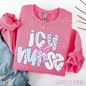 May include: A Safety Pink sweatshirt with the words "ICU Nurse" in a decorative font. The letters are filled with floral and striped patterns. A pink bow is between the letters "U" and "R". A silver necklace is on top of the sweatshirt.