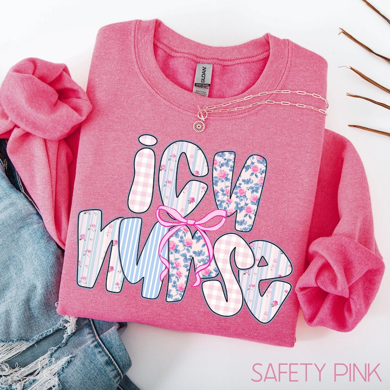 May include: A Safety Pink sweatshirt with the words "ICU Nurse" in a decorative font. The letters are filled with floral and striped patterns. A pink bow is between the letters "U" and "R". A silver necklace is on top of the sweatshirt.
