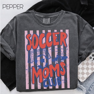 Puede incluir: Sudadera gris oscuro con un dise&ntilde;o gr&aacute;fico. El dise&ntilde;o presenta las palabras "SOCCER MOMS CLUB" en rojo y azul, sobre un fondo de rayas rosas y blancas. Tambi&eacute;n se incluye un bal&oacute;n de f&uacute;tbol y una porter&iacute;a en el dise&ntilde;o.