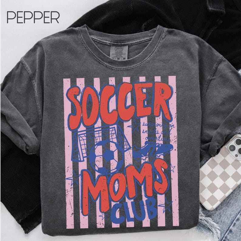 Puede incluir: Sudadera gris oscuro con un dise&ntilde;o gr&aacute;fico. El dise&ntilde;o presenta las palabras "SOCCER MOMS CLUB" en rojo y azul, sobre un fondo de rayas rosas y blancas. Tambi&eacute;n se incluye un bal&oacute;n de f&uacute;tbol y una porter&iacute;a en el dise&ntilde;o.