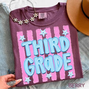 May include: Berry-colored t-shirt with the words "THIRD GRADE" in light blue, outlined in a darker blue, and set against pink vertical stripes. Small star accents are scattered around the text. The shirt is a Comfort Colors XL.
