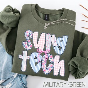 May include: An olive green sweatshirt with the words "SUNY TECH" in a playful font. The letters feature floral, gingham, and striped patterns in pink and blue. A silver necklace with a small pendant rests on the sweatshirt. The text "MILITARY GREEN" is at the bottom.