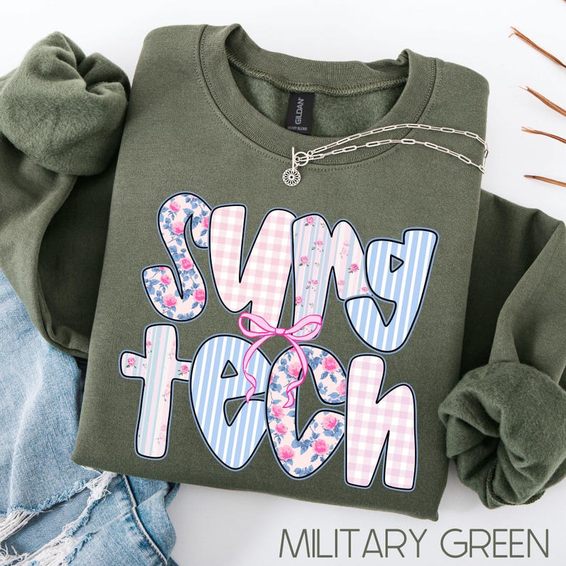 May include: An olive green sweatshirt with the words "SUNY TECH" in a playful font. The letters feature floral, gingham, and striped patterns in pink and blue. A silver necklace with a small pendant rests on the sweatshirt. The text "MILITARY GREEN" is at the bottom.