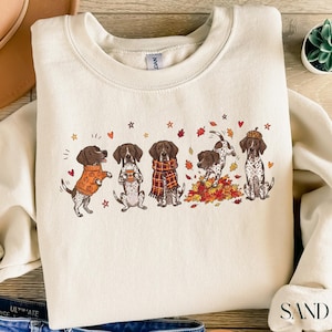 May include: A cream-colored crewneck sweatshirt featuring a graphic print of five spotted dogs in autumn-themed attire. The dogs are depicted wearing sweaters, scarves, and an acorn hat, playing in colorful fall leaves, and holding a coffee cup.