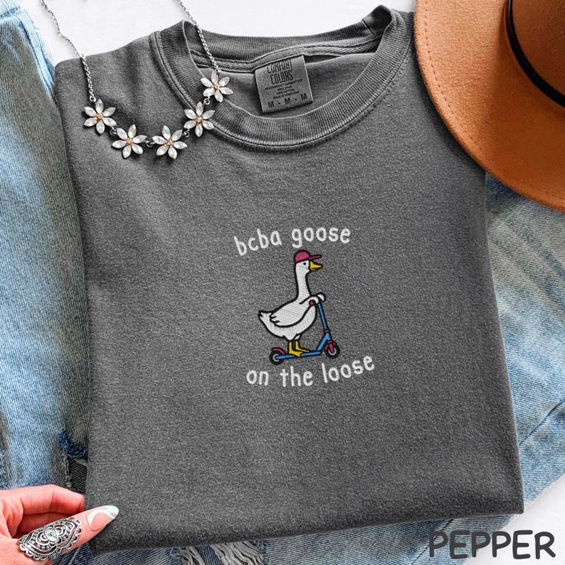 May include: Dark gray t-shirt with the text "bcba goose on the loose" and a cartoon goose riding a scooter. The goose wears a pink cap. A silver necklace with flower-shaped pendants is visible.