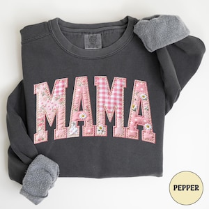 May include: Dark grey sweatshirt with the word "MAMA" in pink patchwork letters. The letters feature floral, gingham, and polka dot patterns. The sweatshirt has grey fleece cuffs and the word "PEPPER" in a circle.
