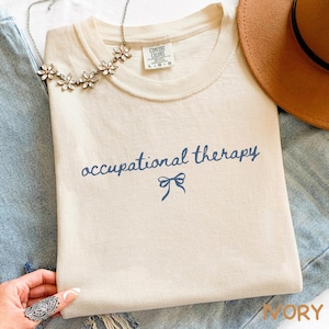 May include: Ivory-colored t-shirt with the words "occupational therapy" in navy blue script, a navy blue bow graphic, and a Comfort Colors tag. A floral necklace and a brown hat are also visible.