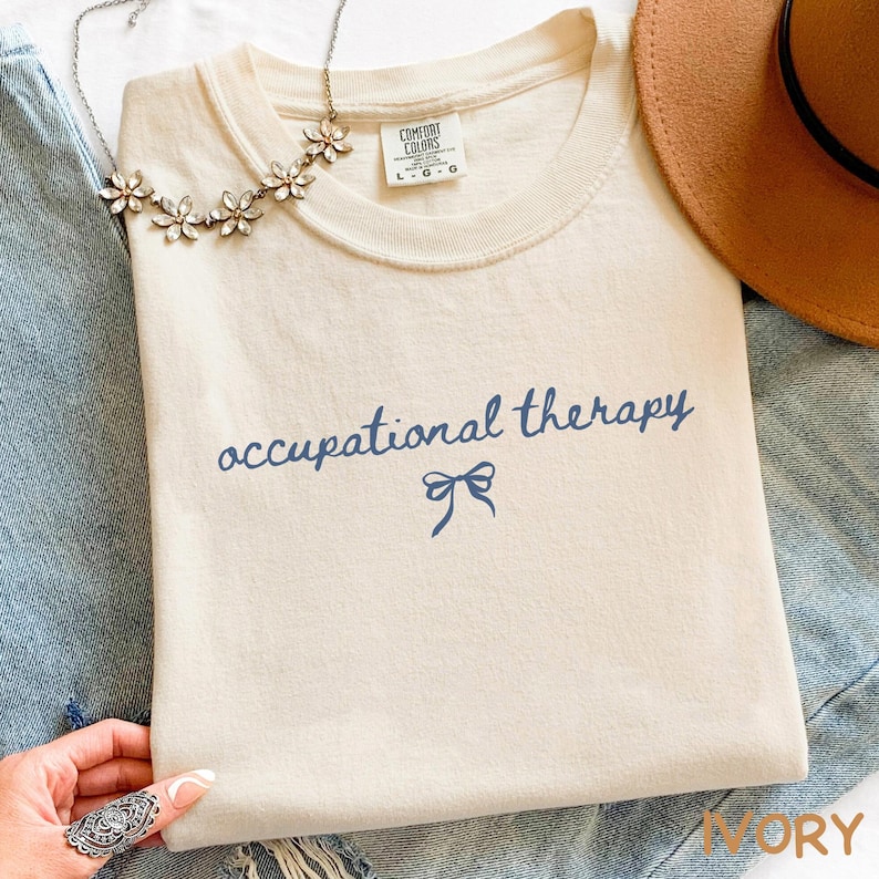 May include: Ivory-colored t-shirt with the words "occupational therapy" in navy blue script, a navy blue bow graphic, and a Comfort Colors tag. A floral necklace and a brown hat are also visible.