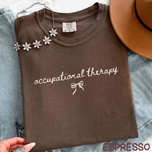 May include: Espresso-colored t-shirt with the words "occupational therapy" in white script and a bow graphic. A floral necklace and a brown hat are also visible. The shirt is folded on top of blue denim.