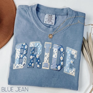 May include: A blue jean colored Comfort Colors t-shirt with the word "BRIDE" appliqued across the front in a patchwork design of floral and gingham patterns. A gold necklace with a pendant rests on the shirt.