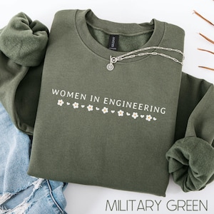 May include: A green sweatshirt with the text "Women in Engineering" and a row of small white flowers and hearts. The sweatshirt is on a blue denim background.