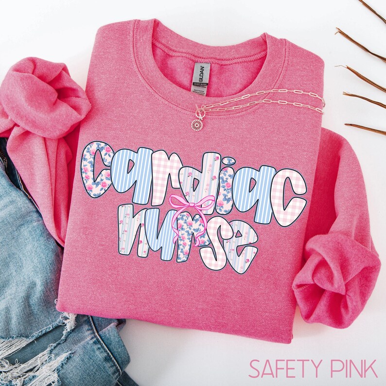 K&ouml;nnte beinhalten: Ein pinkfarbenes Sweatshirt mit dem Schriftzug "Cardiac Nurse". Die Buchstaben sind mit blauen Streifen, floralen Mustern und rosa Vichy-Mustern verziert. Eine Halskette mit Anh&auml;nger liegt auf dem Sweatshirt.