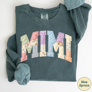 May include: A teal sweatshirt with the word "MIMI" appliqued across the chest in a patchwork design. The letters are made of various floral and patterned fabrics. The sweatshirt has long sleeves and a crew neck.
