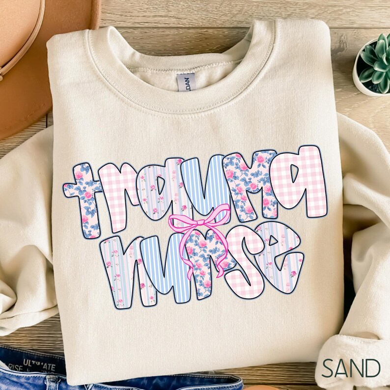 Puede incluir: Una sudadera color arena con las palabras "Trauma Nurse" en una fuente caprichosa. Las letras est&aacute;n decoradas con cuadros rosas, rayas azules y estampados florales, con un lazo rosa. La palabra "SAND" est&aacute; impresa en la esquina inferior derecha.
