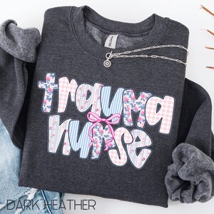 Puede incluir: Sudadera gris oscuro con el texto "Trauma Nurse" en un dise&ntilde;o floral y a rayas. Un lazo rosa est&aacute; entre las palabras. Un collar plateado con un colgante de sol.