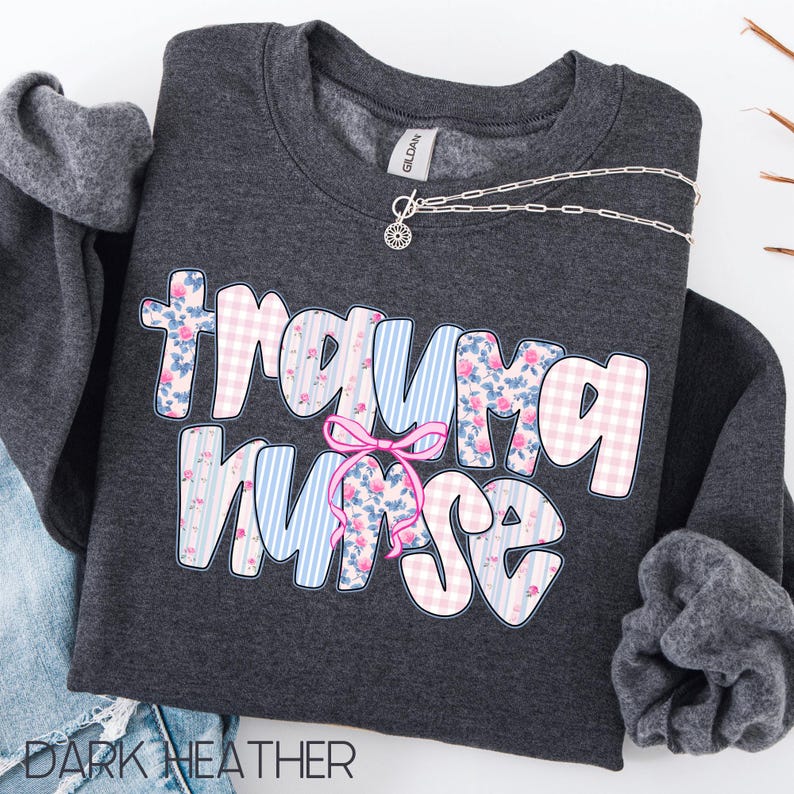 Puede incluir: Sudadera gris oscuro con el texto "Trauma Nurse" en un dise&ntilde;o floral y a rayas. Un lazo rosa est&aacute; entre las palabras. Un collar plateado con un colgante de sol.