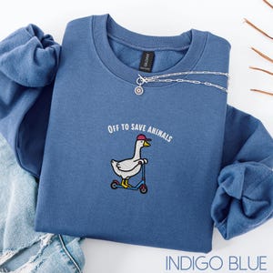 May include: Indigo blue sweatshirt featuring the embroidered text "Off to Save Animals" and a cartoon goose on a scooter, wearing a red cap. A silver necklace with a sun pendant is draped across the sweatshirt.