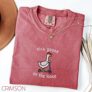 Peut inclure: T-shirt couleur pourpre avec l'inscription "nicu goose on the loose" et une broderie d'une oie sur un scooter. Le t-shirt a un col rond et une petite &eacute;tiquette au niveau de l'encolure. Un collier en argent avec un pendentif est drap&eacute; sur le t-shirt.