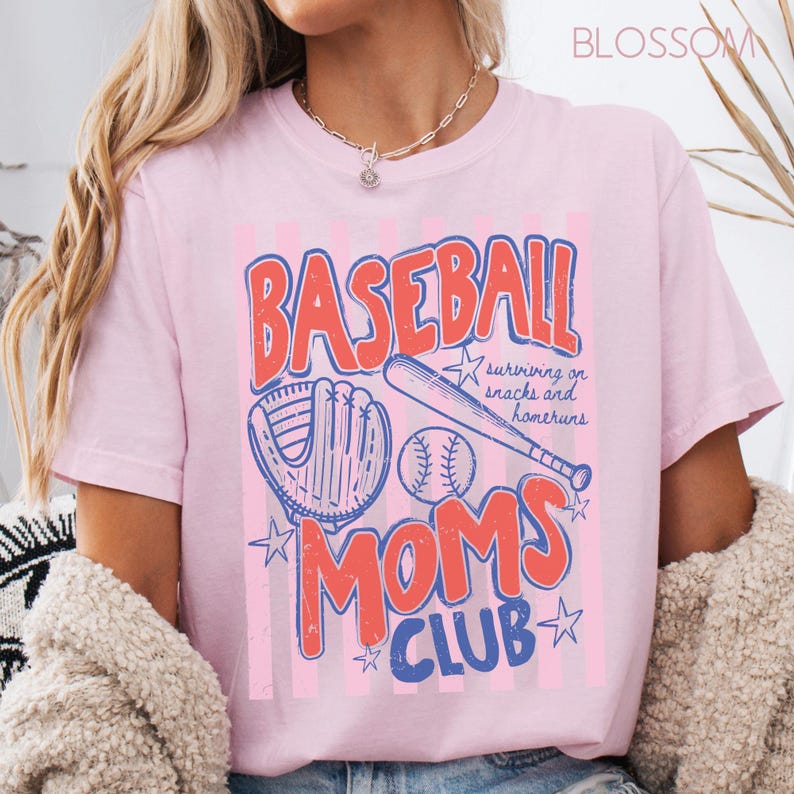Op de afbeelding: Roze T-shirt met een honkbalthema. Het ontwerp bevat de woorden "BASEBALL MOMS CLUB" in rood en blauw, samen met een honkbal, een knuppel en een handschoen. De tekst "surviving on snacks and homeruns" is ook aanwezig.