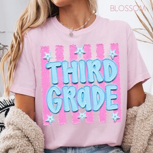 May include: A light pink t-shirt with the words "THIRD GRADE" in light blue, outlined in a darker blue. The text is set against a pink and white striped background with blue star accents. The word "BLOSSOM" is in the top right corner.