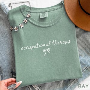 May include: A sage green t-shirt with the words "occupational therapy" in white script and a bow graphic. A silver necklace with flower-shaped accents and a brown hat are also visible. The shirt is folded on top of a pair of blue jeans.