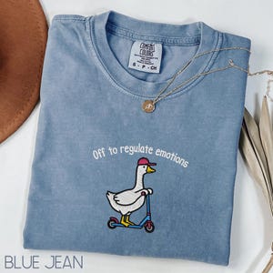May include: A blue jean coloured t-shirt with the text "Off to regulate emotions" and an embroidered goose wearing a baseball cap and riding a scooter. A gold necklace is draped across the shirt. The shirt has a Comfort Colors tag.