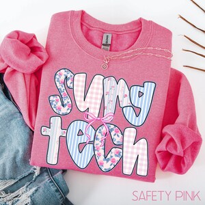 May include: A pink sweatshirt with the words "SUNY TECH" in a playful font. The letters are decorated with floral, gingham, and striped patterns. A pink bow is tied between the words. A delicate necklace rests on the shirt.