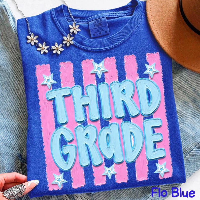 May include: A blue t-shirt with the words "THIRD GRADE" in light blue, set against pink vertical stripes. White star accents are scattered around the text. A floral necklace and a brown hat are visible.