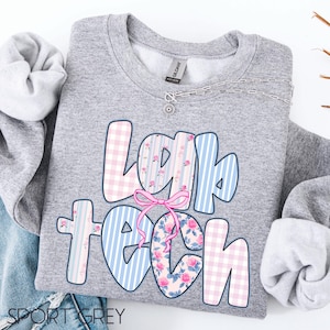 May include: A grey sweatshirt featuring the text "Lap Tech" in a colourful design. The letters are decorated with pink gingham, blue stripes, and floral patterns. A pink bow is centered below the word "Lap". A silver necklace is visible.