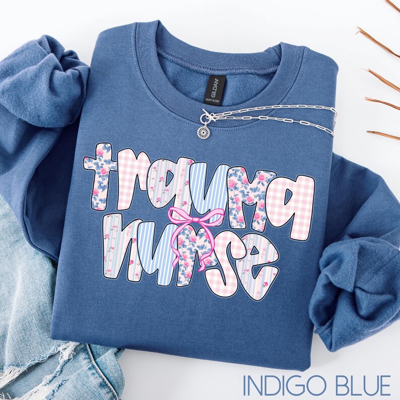 Puede incluir: Sudadera azul &iacute;ndigo con la frase "Trauma Nurse" en una fuente l&uacute;dica. Las letras est&aacute;n decoradas con patrones florales, cuadros y rayas. Un collar plateado con un peque&ntilde;o colgante est&aacute; colocado encima. La sudadera est&aacute; doblada sobre una superficie clara.