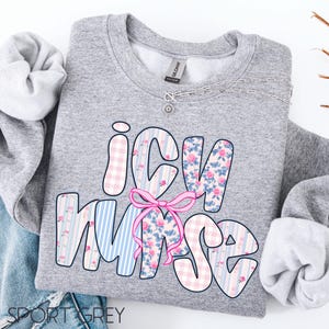 May include: Sport grey sweatshirt with the words "ICU Nurse" in a colorful design. The letters are decorated with pink gingham, blue stripes, and floral patterns, with a pink bow in the center. A silver necklace with a small pendant is also visible.