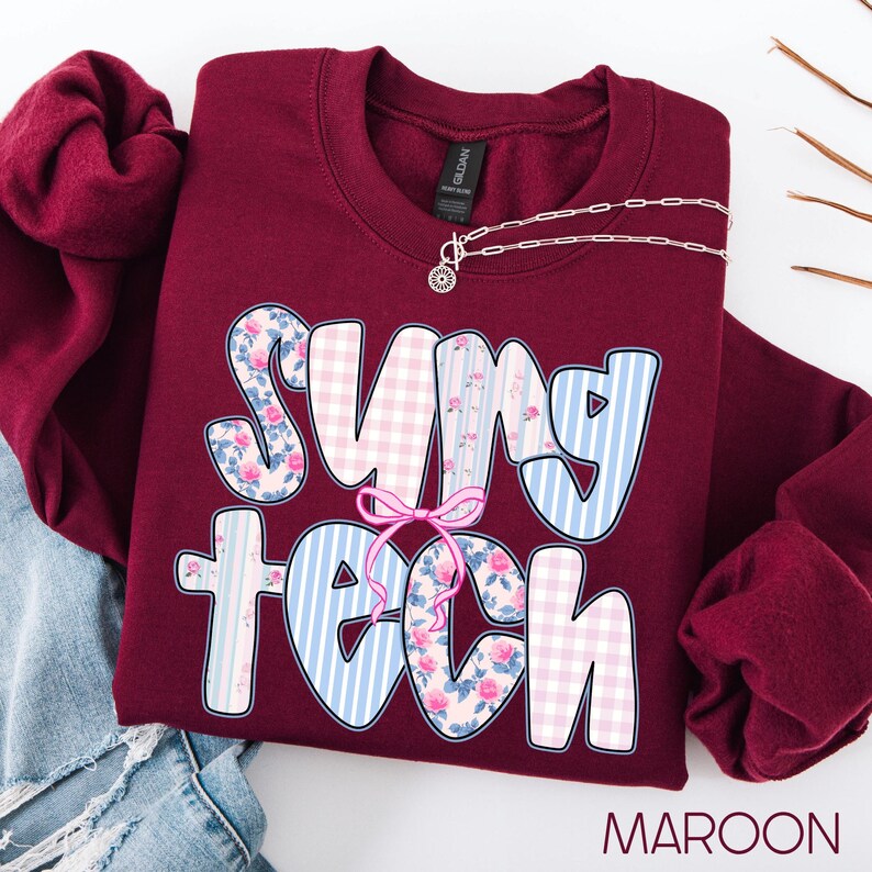 May include: Maroon sweatshirt with the text "Sung Tech" in a playful font. The letters are decorated with floral, gingham, and striped patterns. A silver necklace with a sun pendant rests on the sweatshirt.