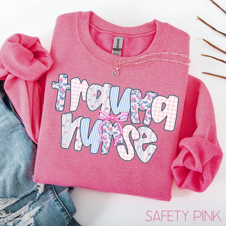 Puede incluir: Una sudadera rosa con la frase "Trauma Nurse" en una fuente l&uacute;dica. Las letras est&aacute;n decoradas con patrones florales, cuadros y rayas. Un lazo rosa est&aacute; colocado entre las palabras. Un collar plateado con un colgante est&aacute; sobre la camisa.