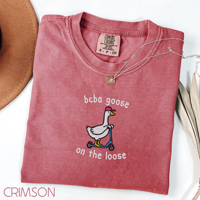 May include: Crimson-colored t-shirt with the text "bcba goose on the loose" and an embroidered goose on a scooter. The shirt has a crew neck and short sleeves. A gold necklace is draped across the shirt.