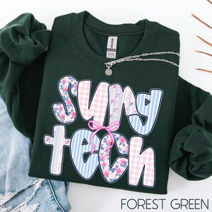May include: Forest green sweatshirt with the text "SUNY TECH" in a playful font. The letters feature floral, gingham, and striped patterns. A silver necklace with a sun pendant is draped on the sweatshirt.