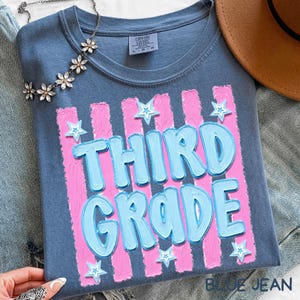 May include: A blue t-shirt with the words "THIRD GRADE" in light blue, outlined in a darker blue, with pink vertical stripes and white star accents. A silver necklace with flower-shaped pendants is draped on the shirt.