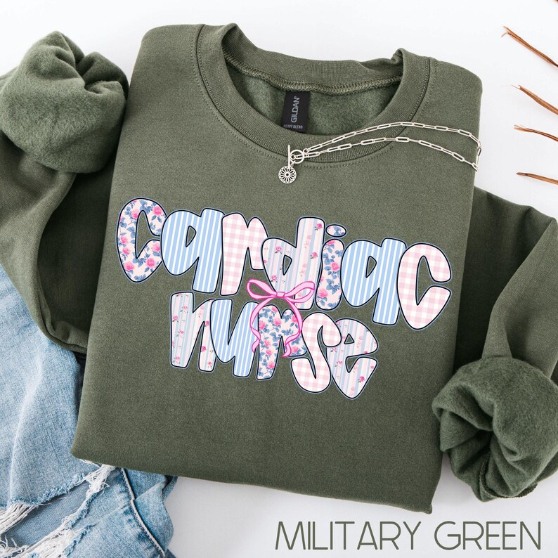 K&ouml;nnte beinhalten: Ein olivgr&uuml;nes Sweatshirt mit dem Schriftzug "Cardiac Nurse" in einer verspielten Schriftart. Die Buchstaben sind mit blauen Streifen, rosa Vichy-Karo und floralen Mustern verziert. Eine silberne Halskette mit einem kleinen Anh&auml;nger liegt auf dem Sweatshirt.