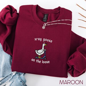 K&ouml;nnte beinhalten: Burgunderfarbenes Sweatshirt mit dem gestickten Schriftzug "xray goose on the loose" und einer Gans auf einem Roller. Eine silberne Halskette mit einem Sonnenanh&auml;nger liegt auf dem Sweatshirt. Das Wort "MAROON" steht unten im Bild.
